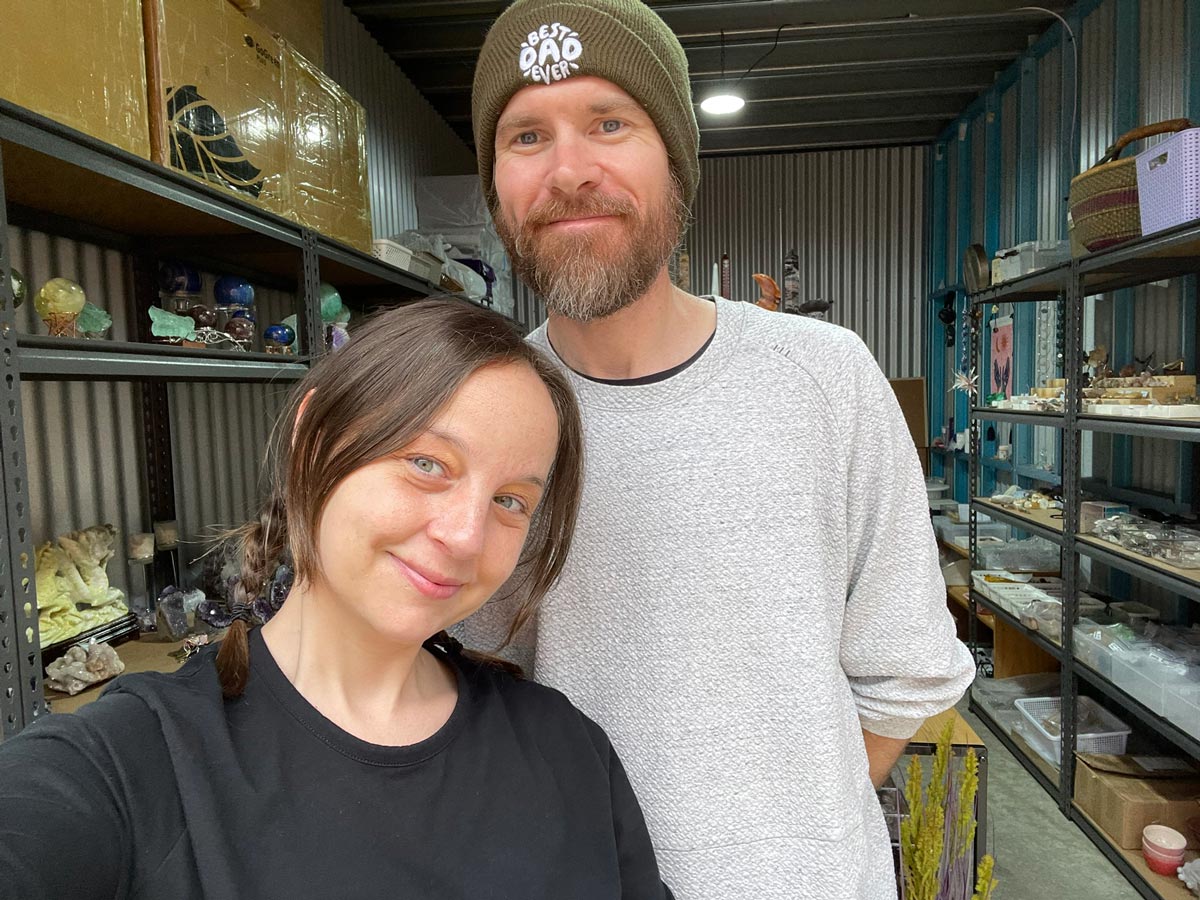 A selfie of Rebecca and Ben in a warehouse full of crystals. Rebecca is shorter, with medium-length brown hair in a plait, and Ben is taller, with a brown and grey beard. 
