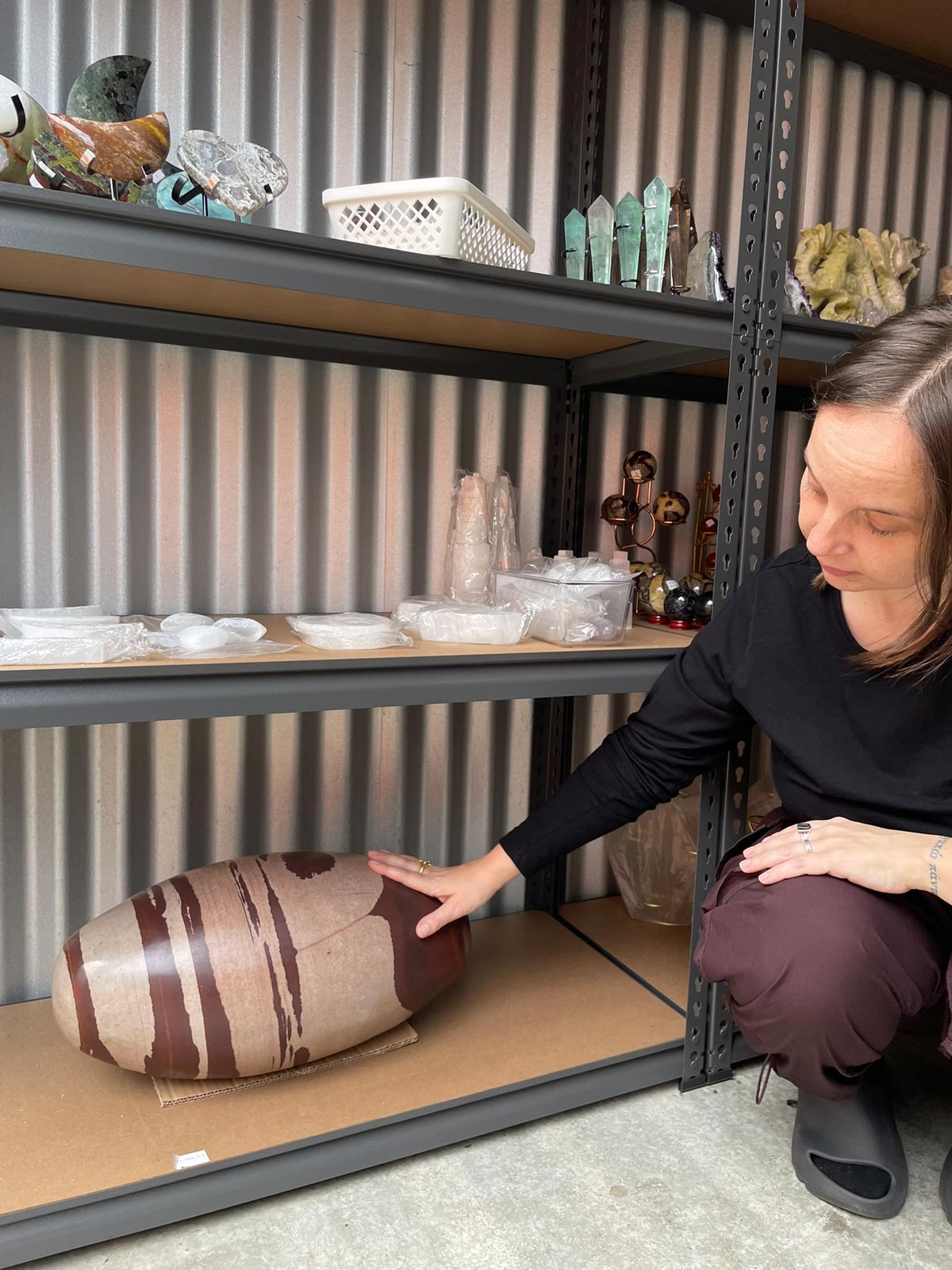 Rebecca kneeling to touch a large piece of jasper on a bottom shelf. The higher shelves are filled with smaller crystals. 